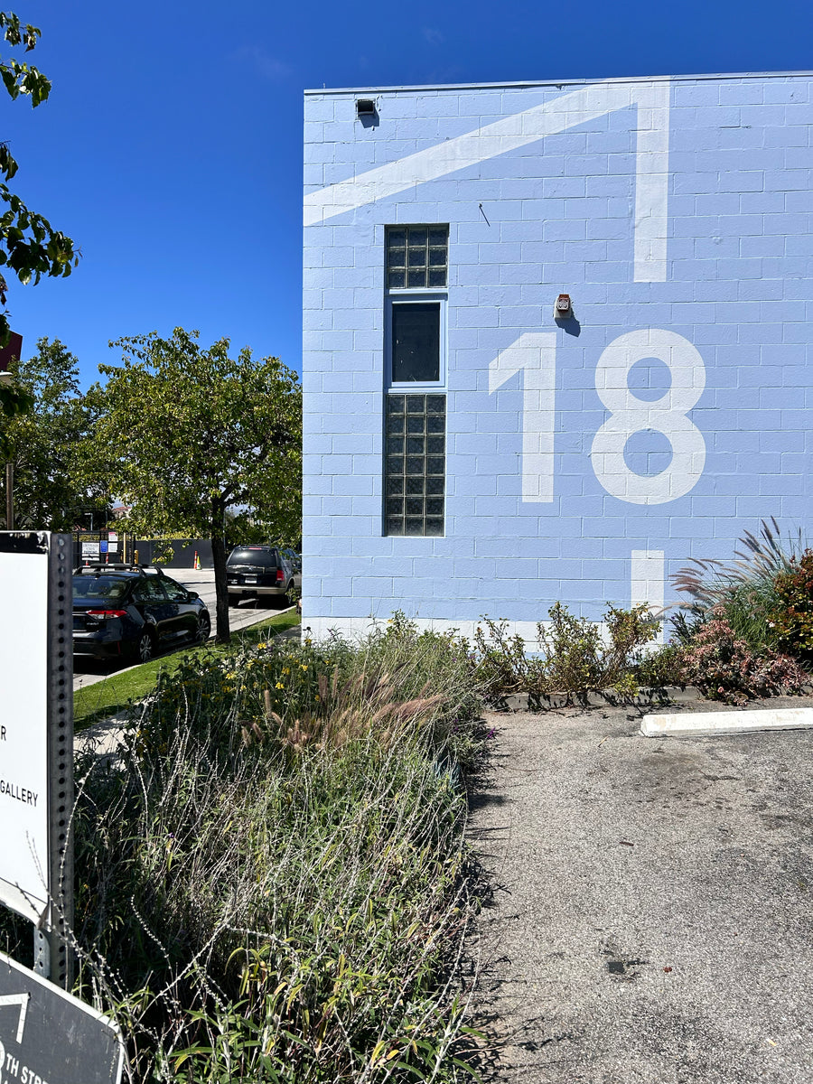 A light blue brick building exterior with large white painted numbers '18' and a window, surrounded by tall grasses, shrubs, parked cars, and a clear blue sky.