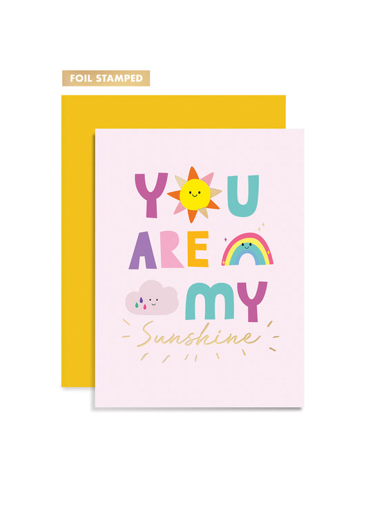 A bright pink greeting card featuring "You are my sunshine" in colorful text and cute illustrations with gold foil accents, paired with a yellow envelope. 