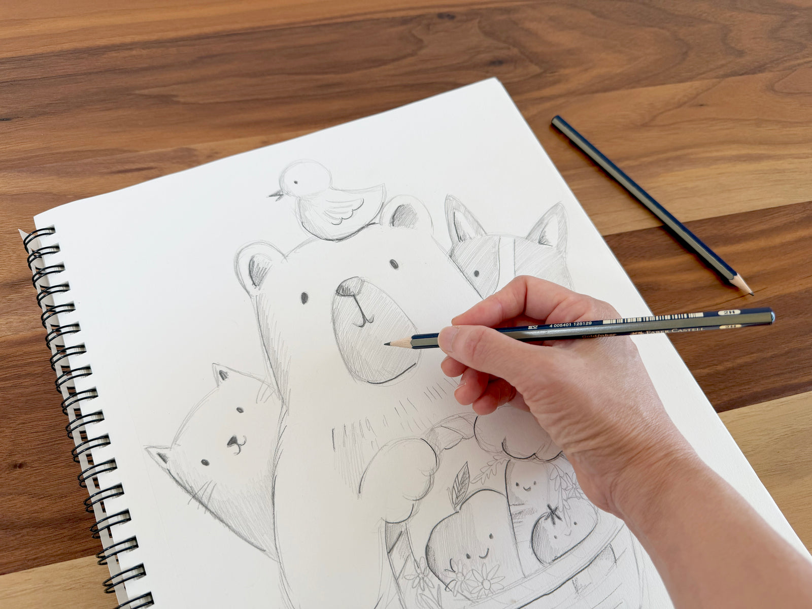 A hand holding a graphite pencil sketches a cute bear, cat, dog, and bird illustration in a spiral-bound sketchbook on a wooden table, with two pencils nearby.