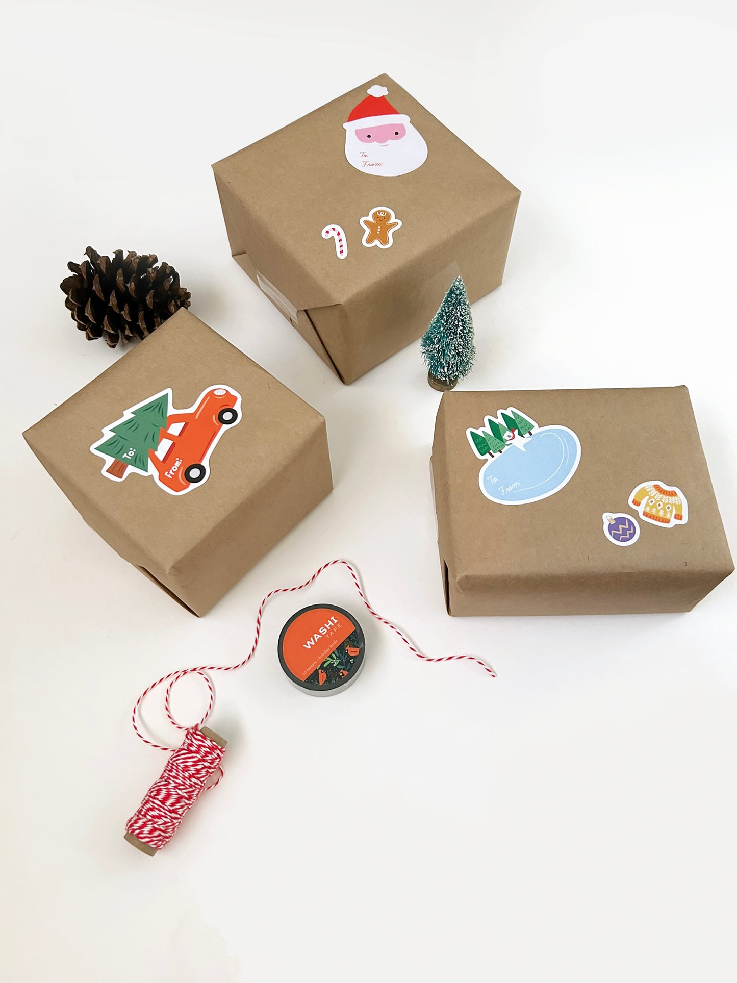 Three small kraft paper gift boxes decorated with holiday gift stickers, arranged on a white background with a pinecone, a miniature tree, and red-and-white twine.