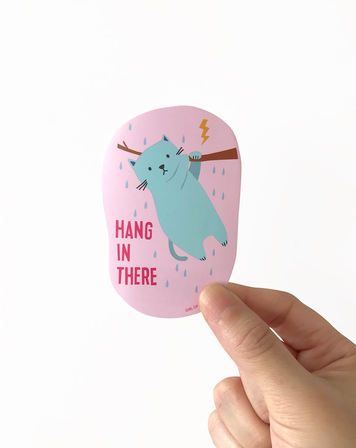 Pink card vinyl sticker with a blue cat illustration and 'Hang in there' text, held by a hand on a white background