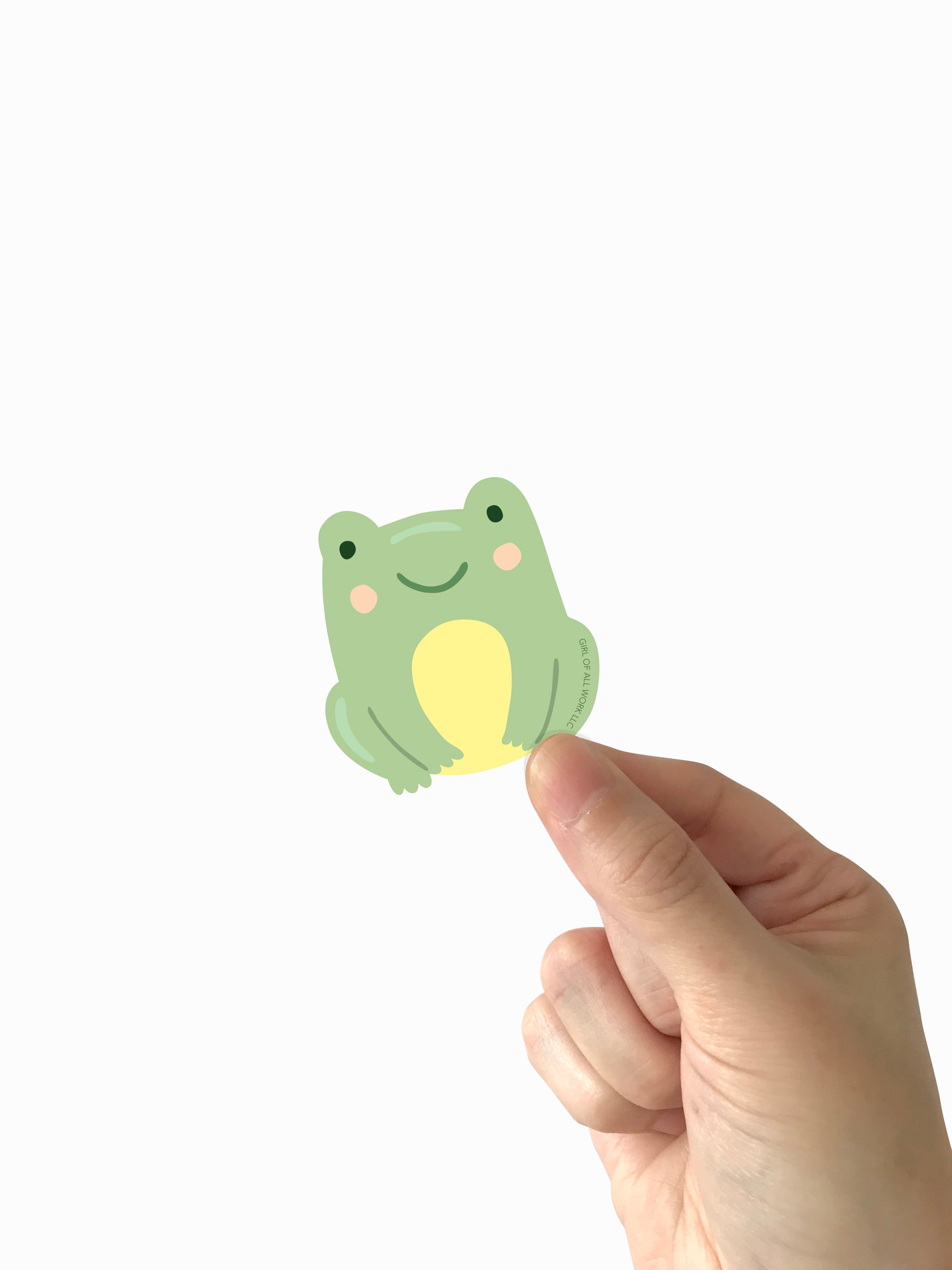 Hand holding a small green frog-shaped vinyl sticker against a white background