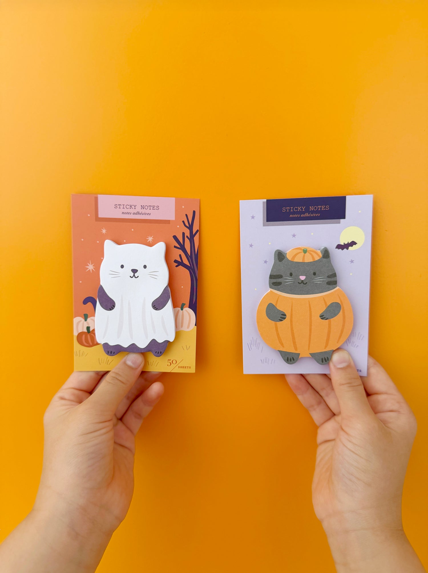 Two hands hold two Halloween-themed sticky notes against a solid orange background. The left sticky note card features a white ghost cat on an orange background, and the right sticky note card features a gray cat in a pumpkin costume on a light purple background.
