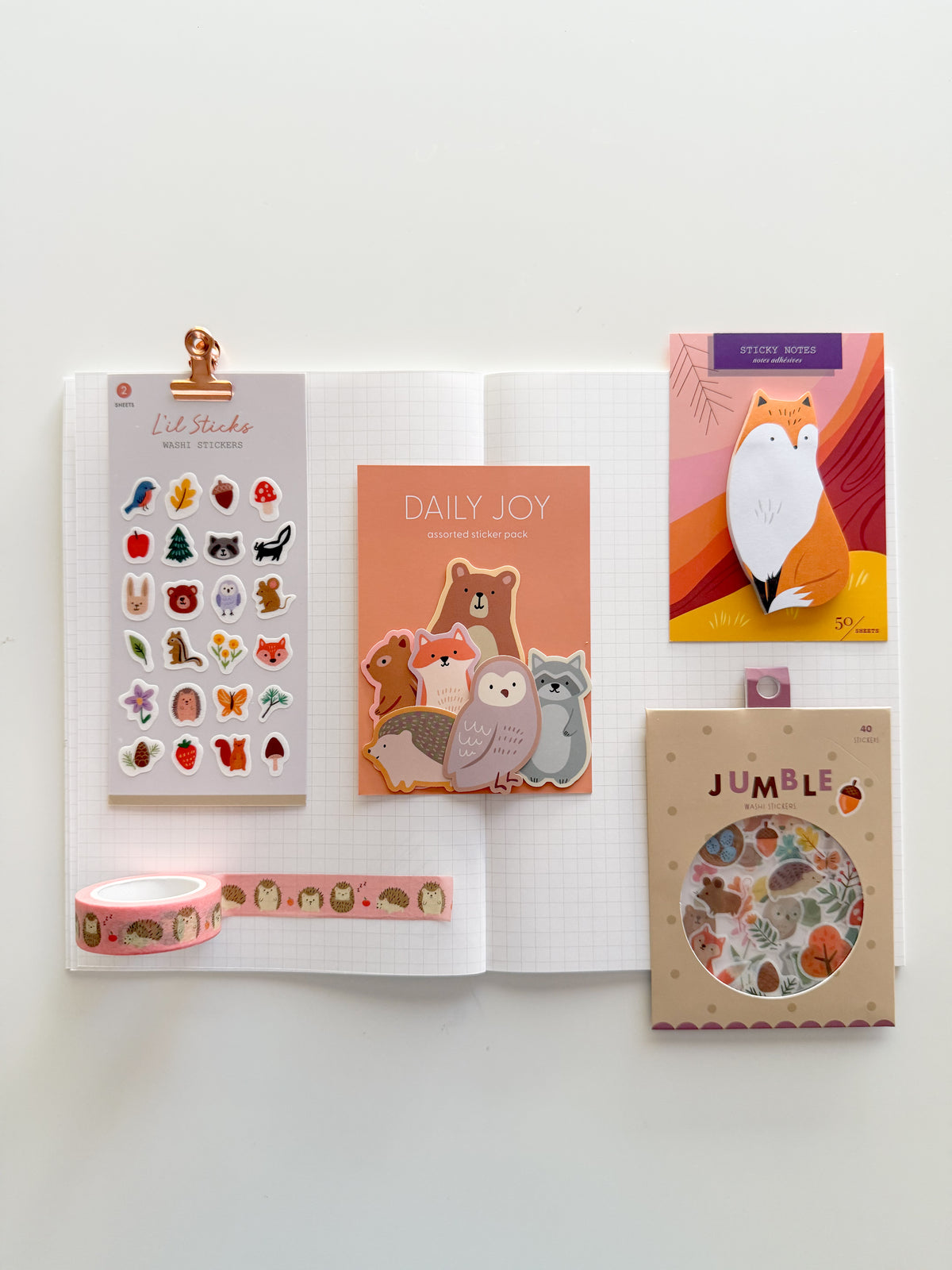 A flat-lay display of cute, woodland-themed stationery items arranged on an open white grid notebook, including a fox die-cut sticky note, Forest Fun L’il Sticks Washi Stickers, Forest Animals Flake Stickers, Woodland Friends Jumble Washi Stickers, and Hedgehog washi tape.