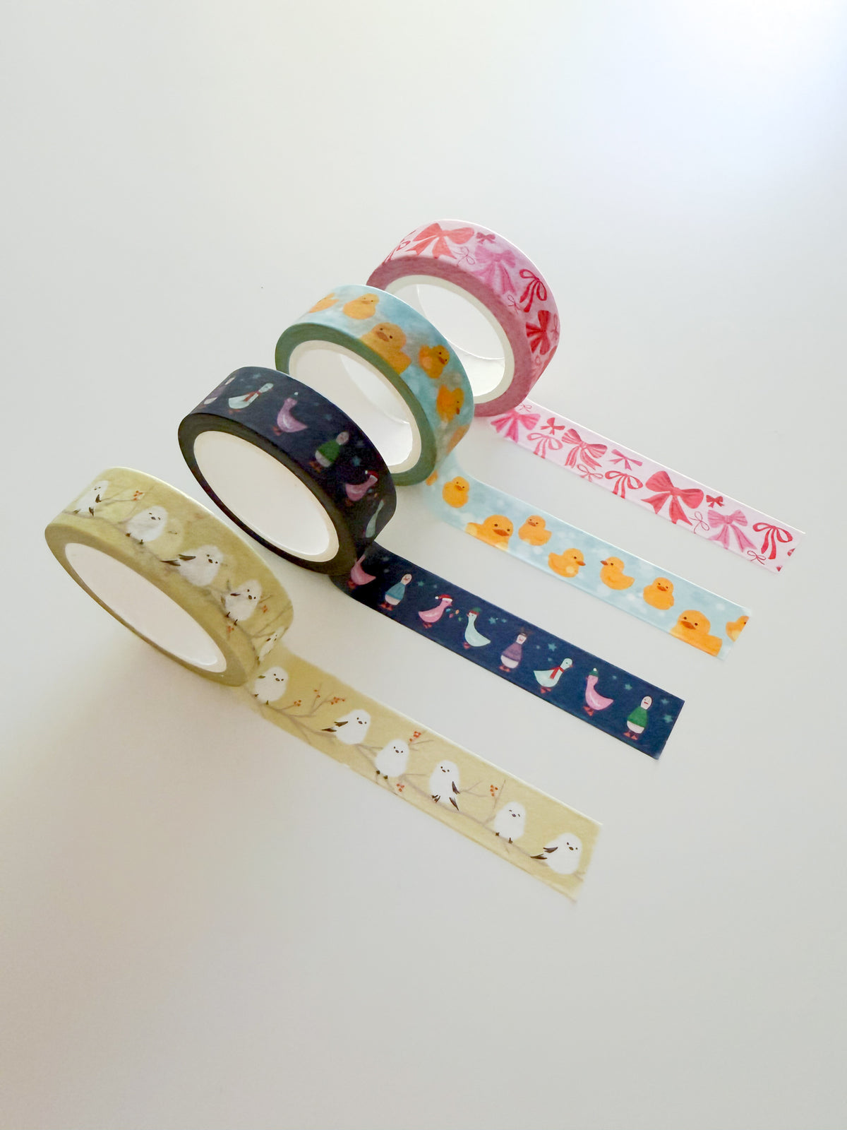 Four different washi tape designs arranged on a white background, with a short length of each tape unrolled to show its pattern. The designs are Snow Fairies Washi Tape, Rubber Duckies Washi Tape, Festive Geese Washi Tape, and Ribbons Washi Tape.